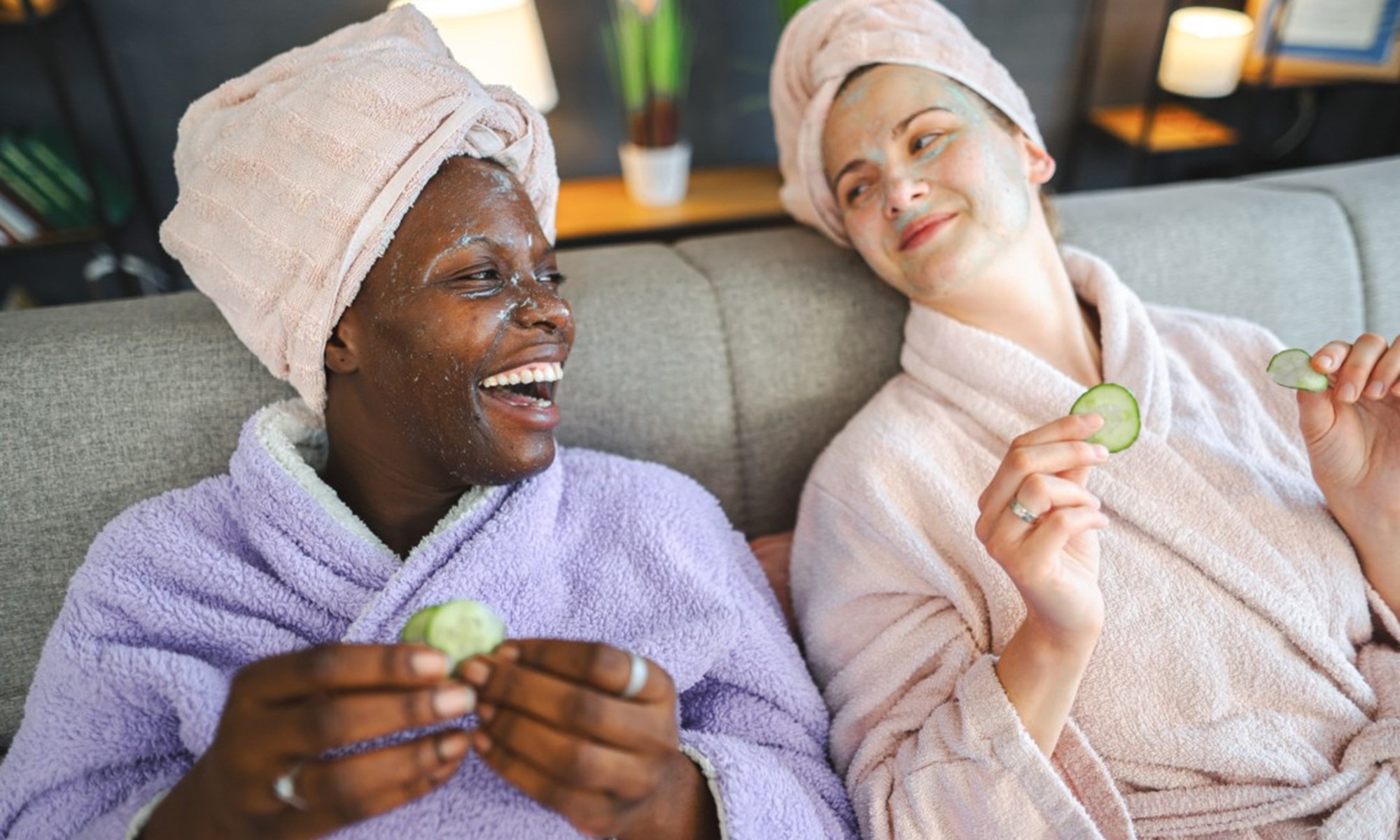 two women in bathrobes with face masks holding cucumber slices
